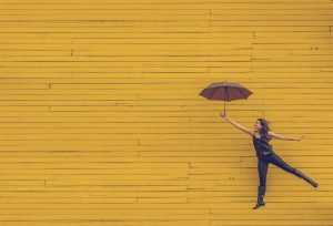 Keltainen puurakennus, jonka alareunassa on tummiin vaatteisiin puketunut nainen, joka hyppää sateenvarjo auki kädessään.